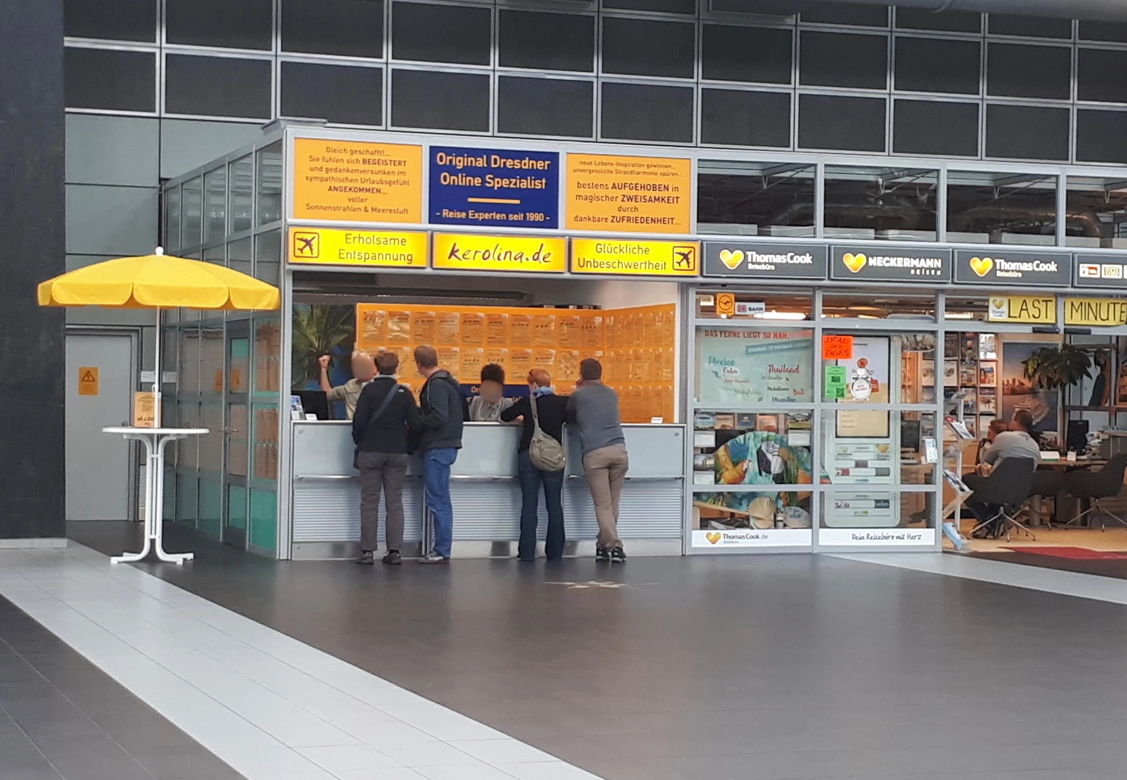KEROLINA Reisebüro-Counter im Flughafen Dresden mit Kunden am Tresen, gelb-blaue Beschilderung mit kerolina.de-Logo, daneben Thomas Cook und Neckermann